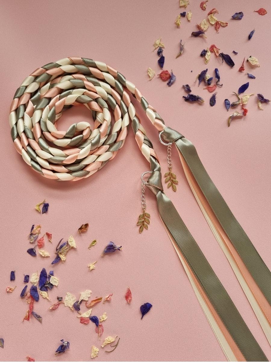Intertwined Handfasting Cord made from cream, sage and peach coloured strands of satin ribbon, along with gold leaf charms. Set against a pink backdrop with confetti.