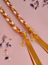 Load image into Gallery viewer, Intertwined Handfasting Cord made from gold, amber and sand coloured strands of satin ribbon, along with gold bee charms. Set against a pink backdrop with confetti.