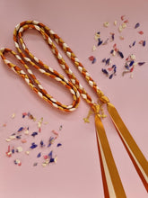 Load image into Gallery viewer, Intertwined Handfasting Cord made from gold, amber and sand coloured strands of satin ribbon, along with gold bee charms. Set against a pink backdrop with confetti.