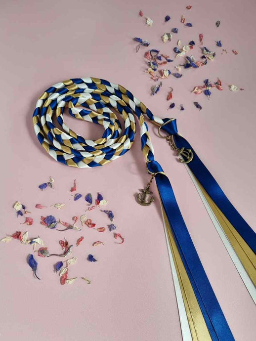 Intertwined Handfasting Cord made from blue, ivory and gold strands of satin ribbon.  Set against a pink backdrop with confetti.