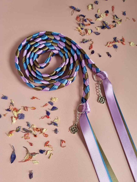 Intertwined Handfasting Cords made from mauve, blue and green strands of satin ribbon, along with silver celtic knot charms.  Set against a pink background with confetti.