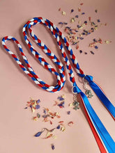 Load image into Gallery viewer, Intertwined Handfasting Cord made from red, white and blue strands of satin ribbon, along with silver rose charms. Set against a pink backdrop with confetti.