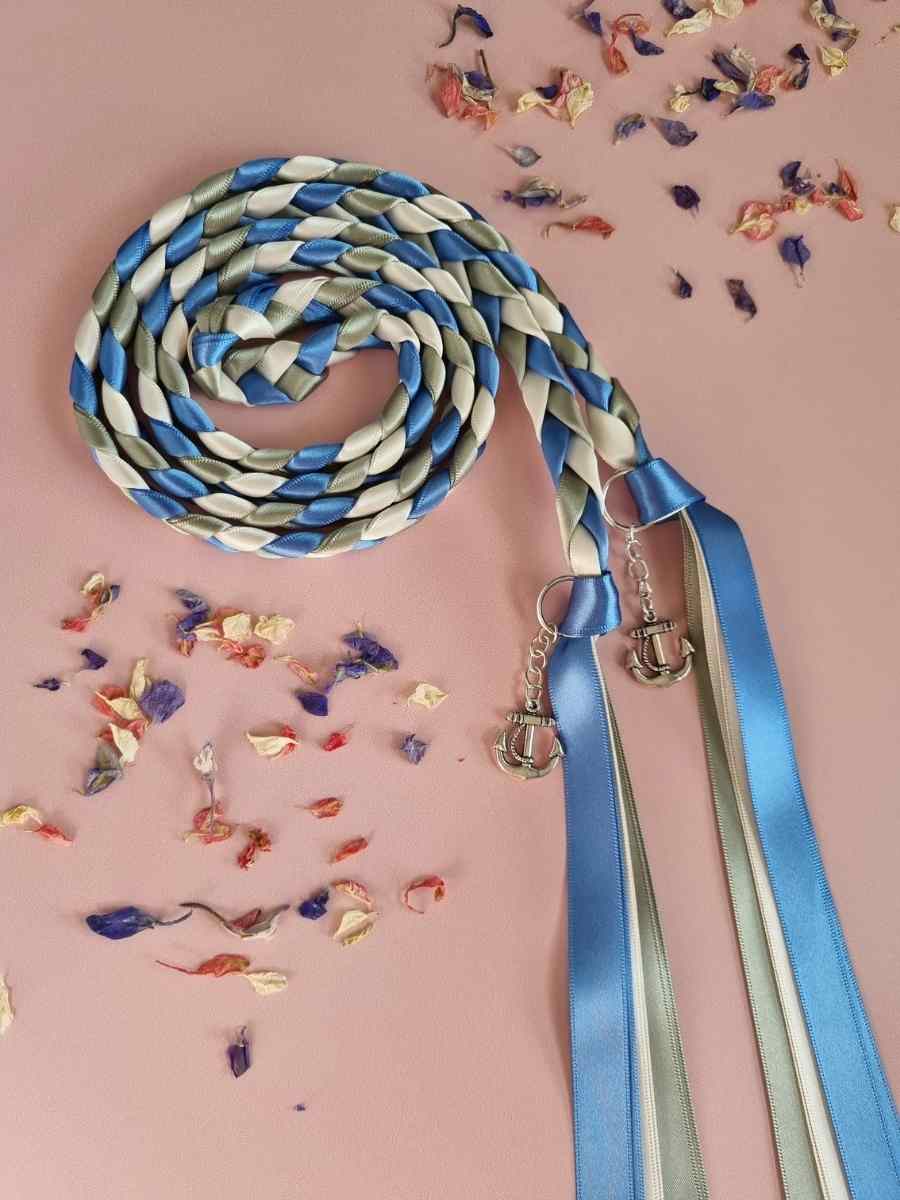 Intertwined Handfasting Cord made from green, blue and ivory strands of satin ribbon, along with silver anchor charms.  Set against a pink backdrop with confetti.