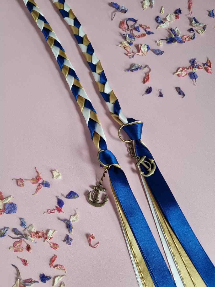 Intertwined Handfasting Cord made from blue, ivory and gold strands of satin ribbon, along with gold anchor charms.  Set against a pink backdrop with confetti.