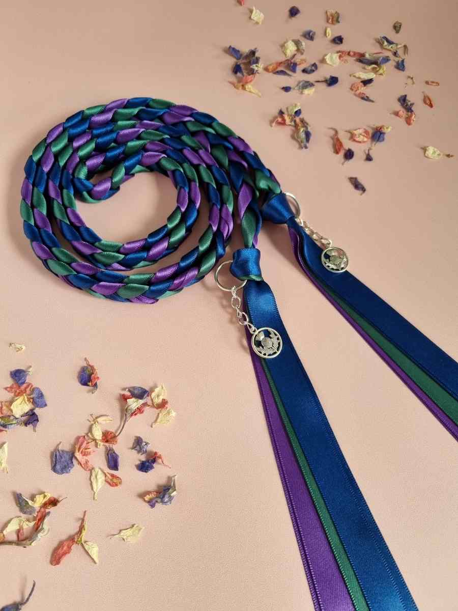 Intertwined Handfasting Cord made from blue, purple and green strands of satin ribbon, along with silver thistle charms.  Set against a pink backdrop with confetti.