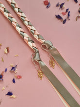 Load image into Gallery viewer, Intertwined Handfasting Cord made from cream, sage and peach coloured strands of satin ribbon, along with gold leaf charms. Set against a pink backdrop with confetti.
