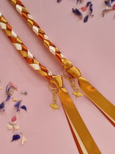 Load image into Gallery viewer, Intertwined Handfasting Cord made from gold, amber and sand coloured strands of satin ribbon, along with gold bee charms. Set against a pink backdrop with confetti.
