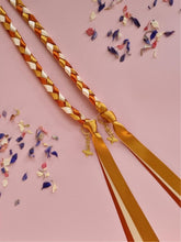 Load image into Gallery viewer, Intertwined Handfasting Cord made from gold, amber and sand coloured strands of satin ribbon, along with gold bee charms. Set against a pink backdrop with confetti.
