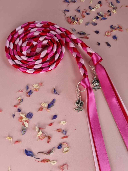 Intertwined Handfasting Cord made from pink, fuchsia and red strands of satin ribbon, along with silver rose charms.  Set against a pink background with confetti.