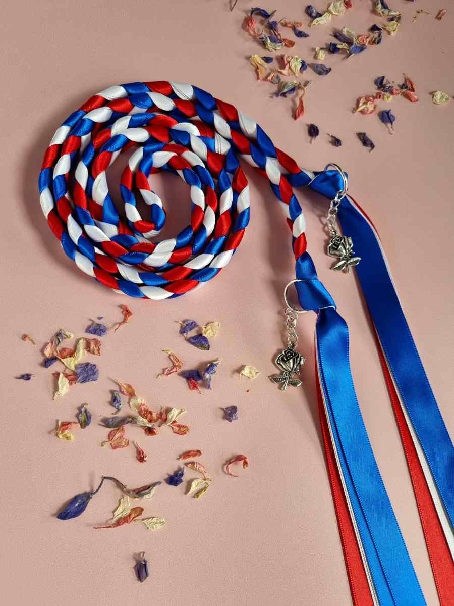 Intertwined Handfasting Cord made from red, white and blue strands of satin ribbon, along with silver rose charms.  Set against a pink backdrop with confetti.